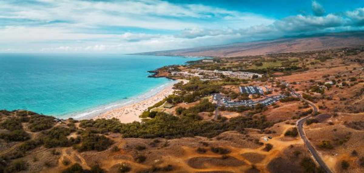 Aerial view of a crescent-shaped sandy beach with turquoise water, resort buildings, and dry hills along the Kohala coastline on Hawaii’s Big Island.