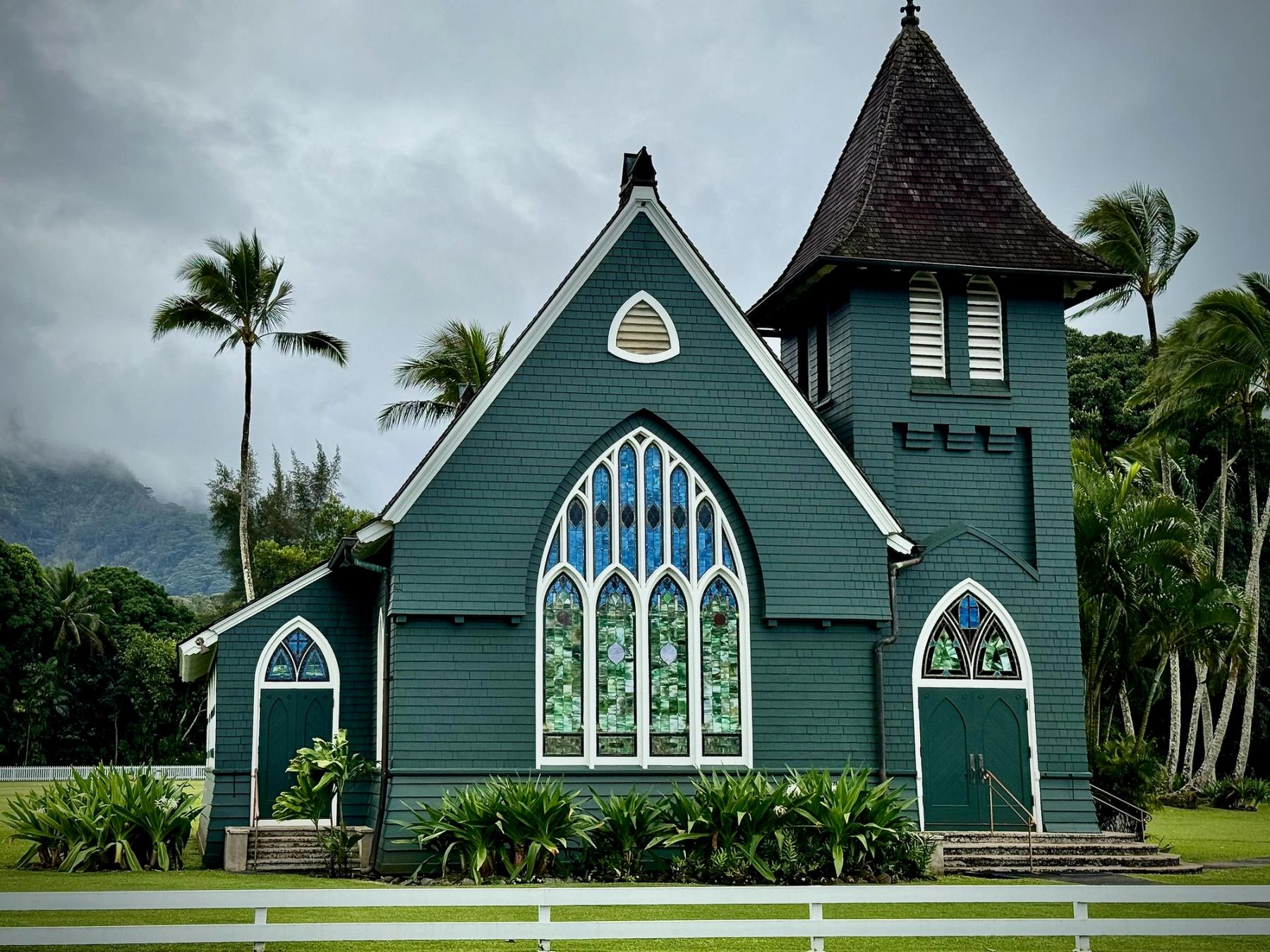 Green wooden church with stained-glass windows, palms, and misty mountains under an overcast sky in Hanalei