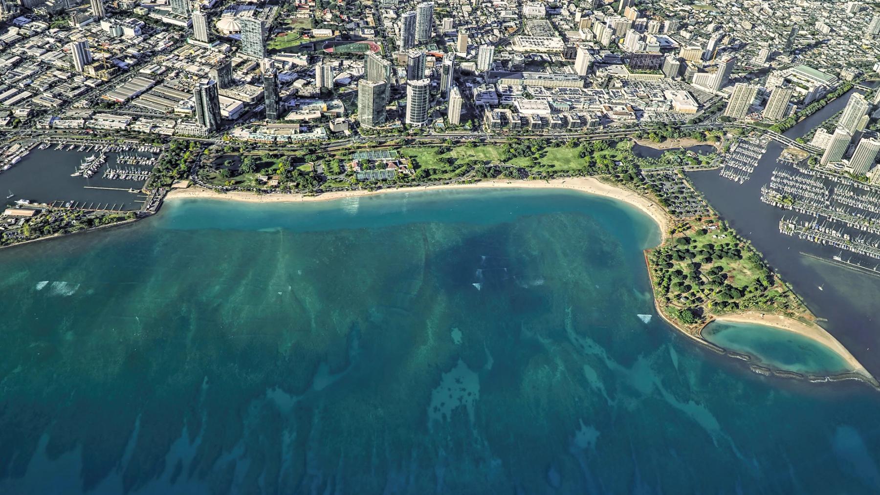 Aerial view of Ala Moana Beach Park in Honolulu, with a sandy shoreline, turquoise water, marinas, and nearby high-rise buildings.