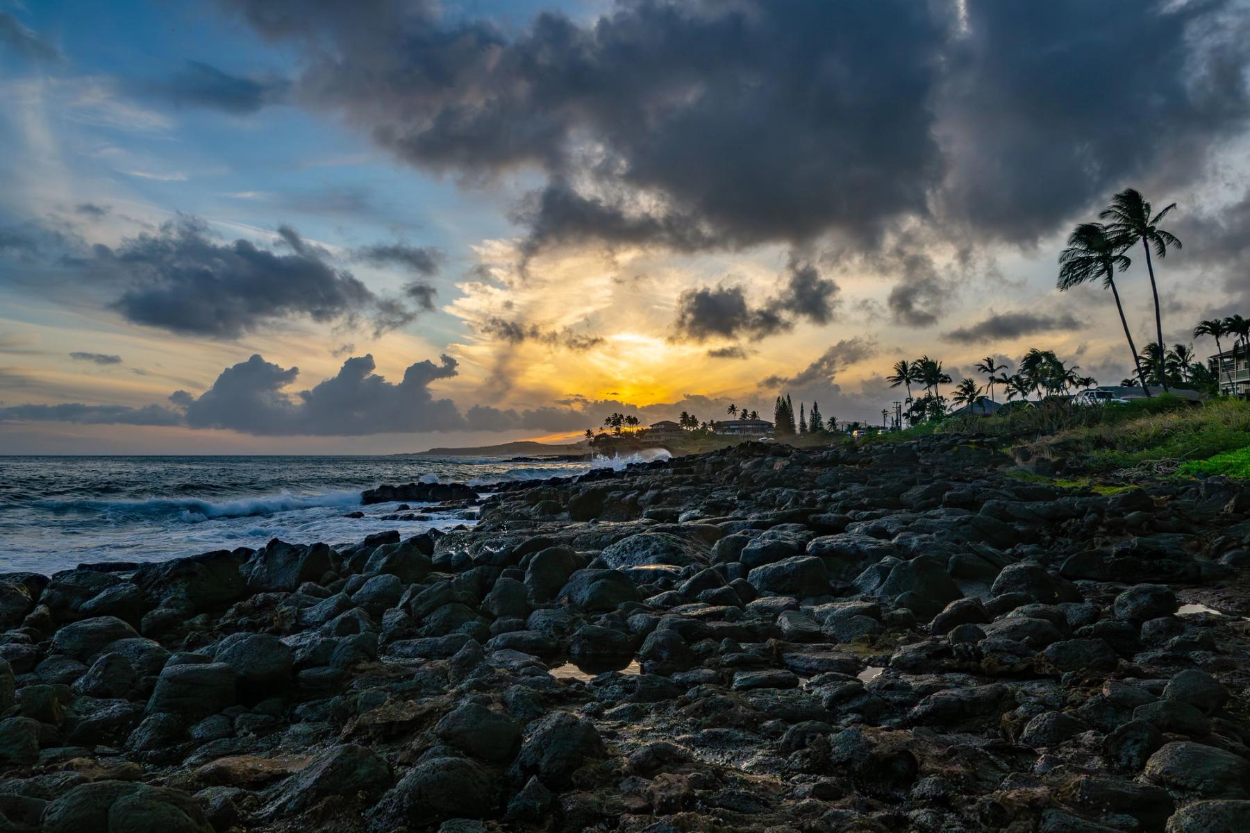 Sunset glow beneath dramatic clouds over a rocky lava shoreline with surf and palm trees along Poʻipū coast on Kauaʻi.