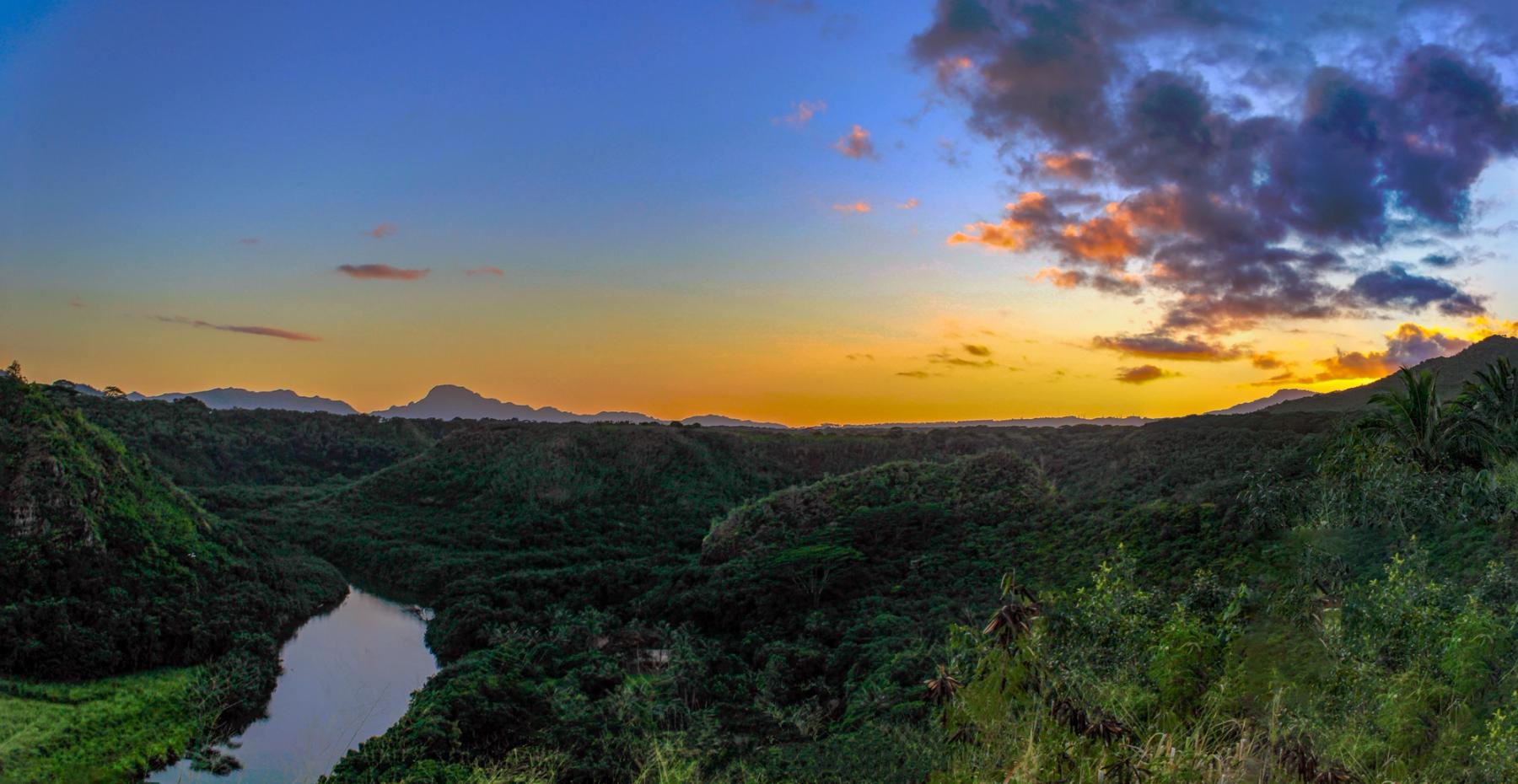 Sunset sky glowing orange and blue above a lush green Kauaʻi valley with a winding river and distant mountain silhouettes