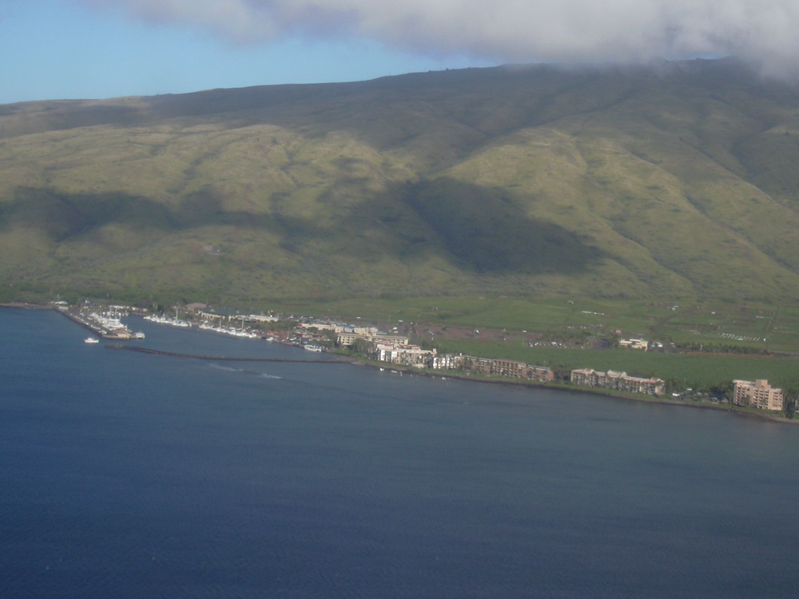 Aerial view of Māʻalaea, Maui, showing a coastal harbor and buildings along the shoreline below green mountains and low clouds.