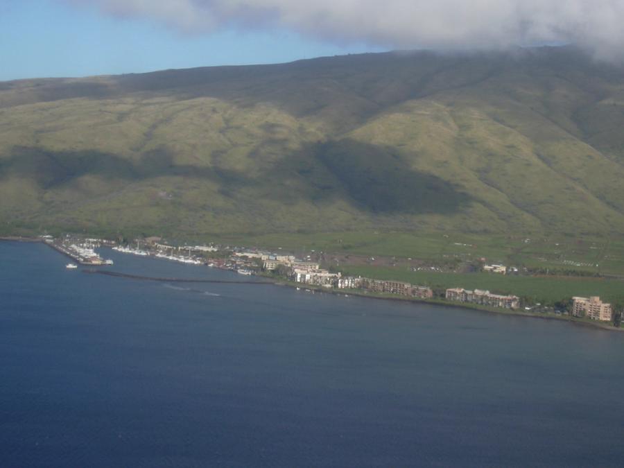 Aerial view of Māʻalaea, Maui, showing a coastal harbor and buildings along the shoreline below green mountains and low clouds.