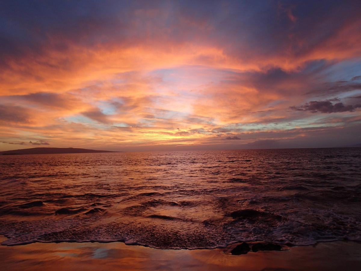 Sunset sky with orange and pink clouds over the ocean, with small waves washing onto a sandy shoreline in South Maui.