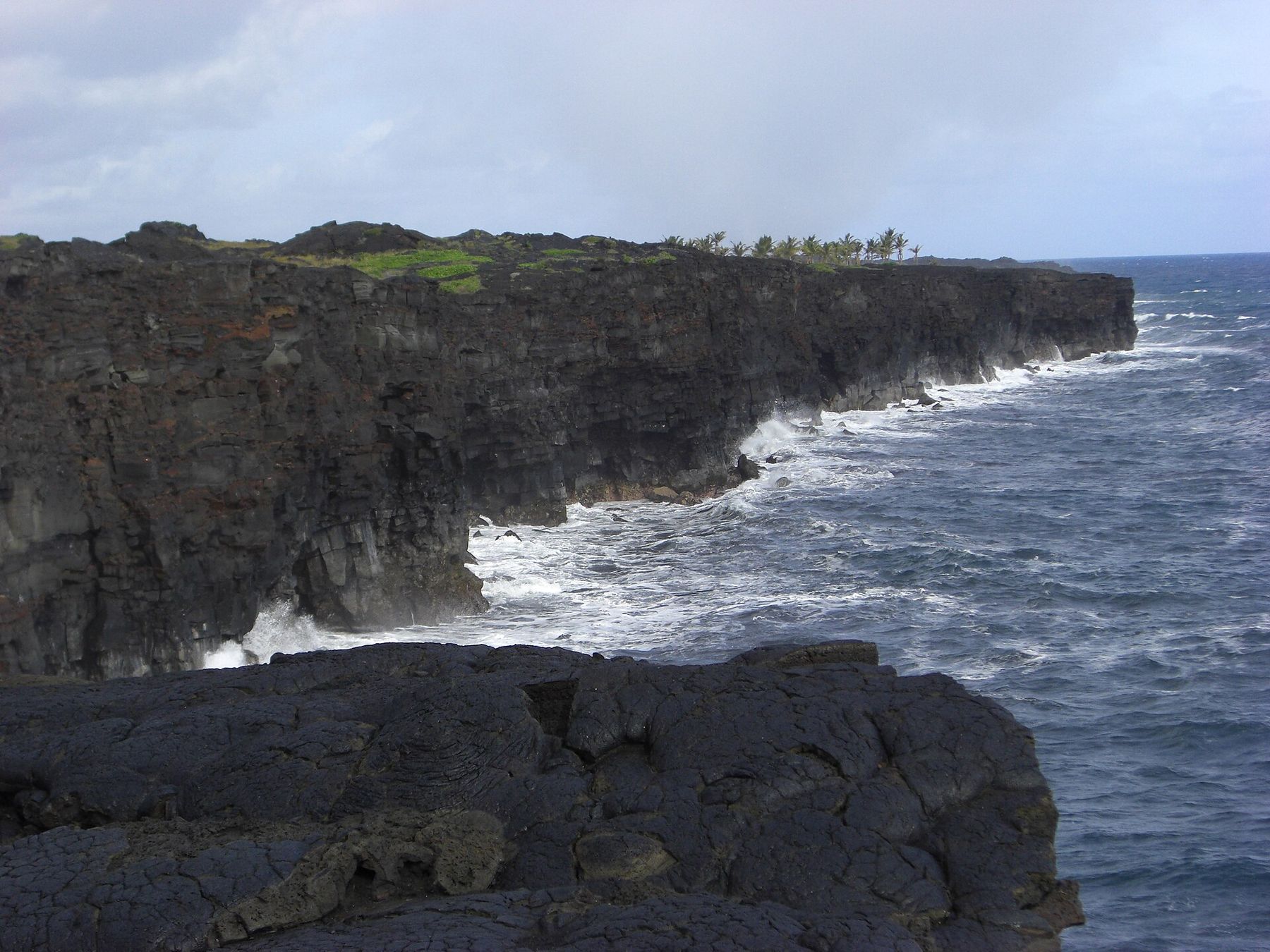 Waves crash against dark lava sea cliffs along the Kalapana coast on Hawaii’s Big Island under an overcast sky.