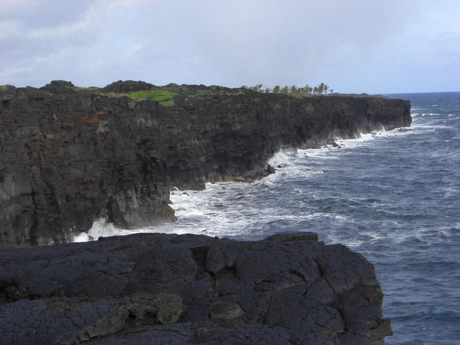 Waves crash against dark lava sea cliffs along the Kalapana coast on Hawaii’s Big Island under an overcast sky.