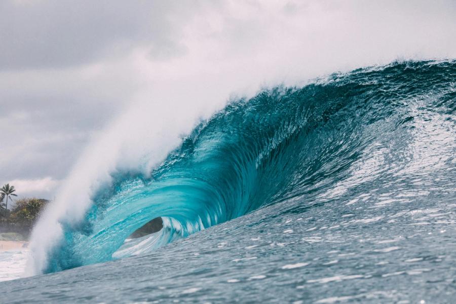 A large turquoise wave curls into a barrel near a sandy shoreline with palm trees beneath a cloudy sky in Pūpūkea, Oʻahu.