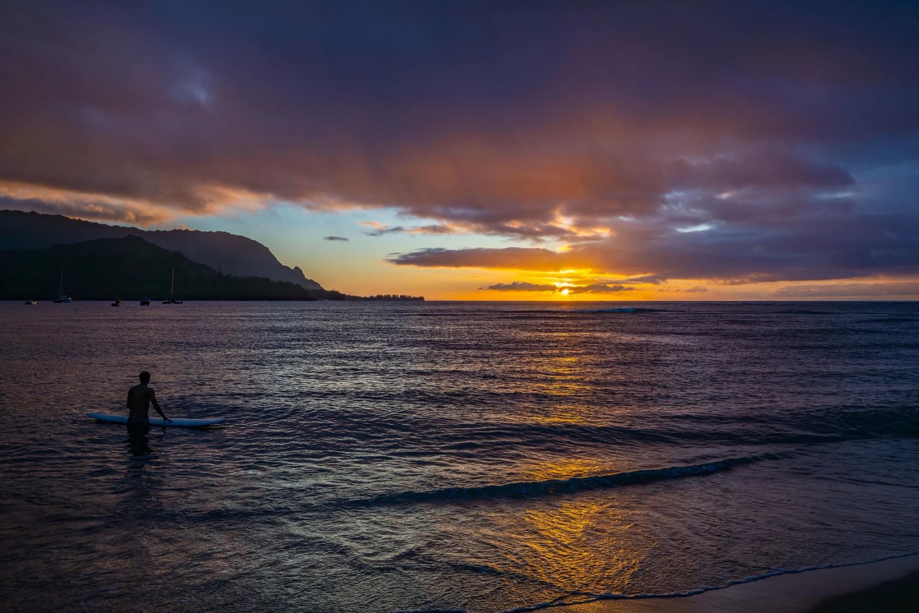 Sun setting over Hanalei Bay with dramatic clouds, golden reflections on the water, a mountain silhouette at left, and a surfer standing beside a board in the shallows.