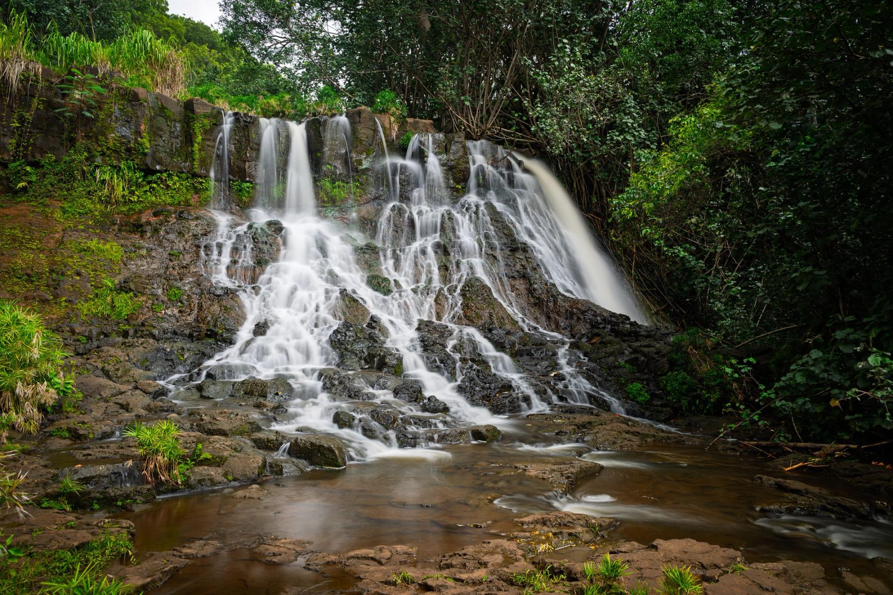 Wide multi-tier waterfall cascading over dark rock into a shallow pool, surrounded by dense green tropical forest