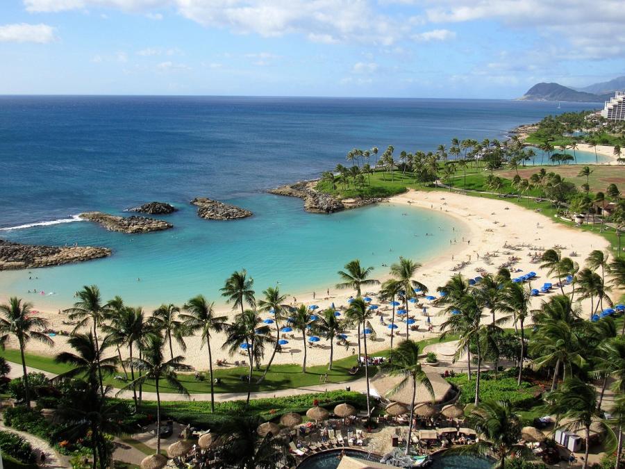 Curved sandy lagoon beach with turquoise water, rock breakwaters, and palm-lined lawns beside resort buildings in Ko Olina, Oahu.