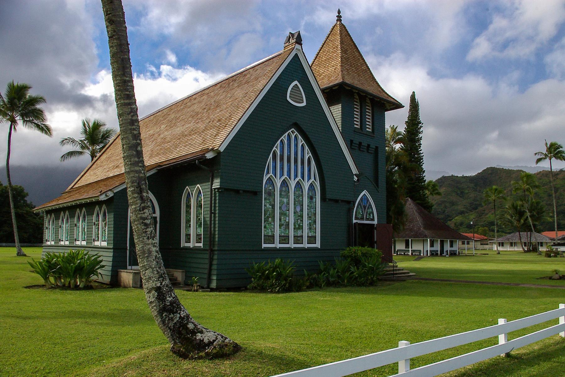 Green wooden church with arched stained-glass windows and a steep roof, framed by palm trees and backed by cloudy mountains in Hanalei