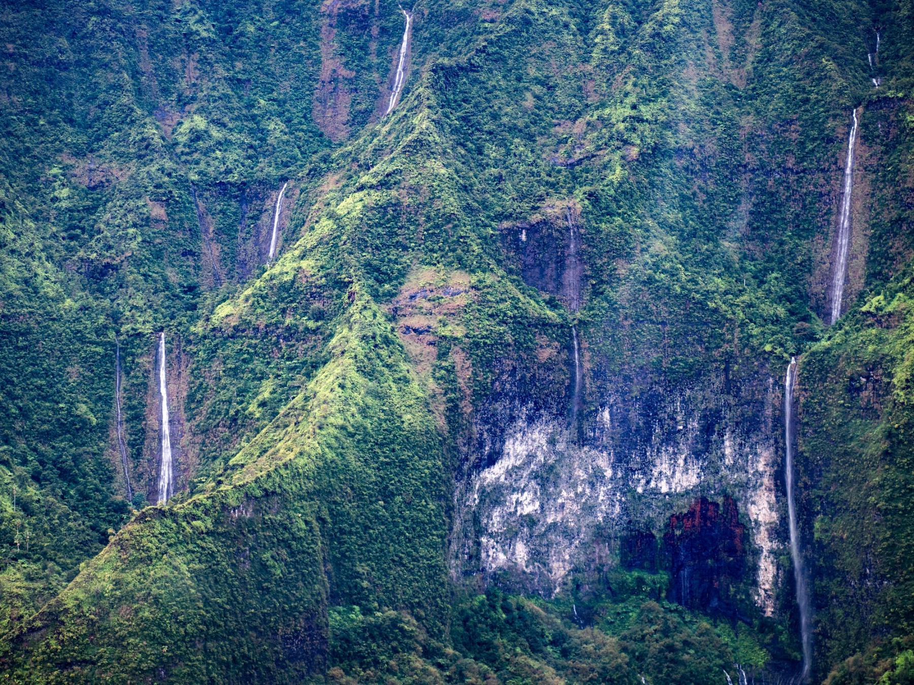 Multiple thin waterfalls streaming down steep, lush green cliffs with dark rock bands and a central ridgeline