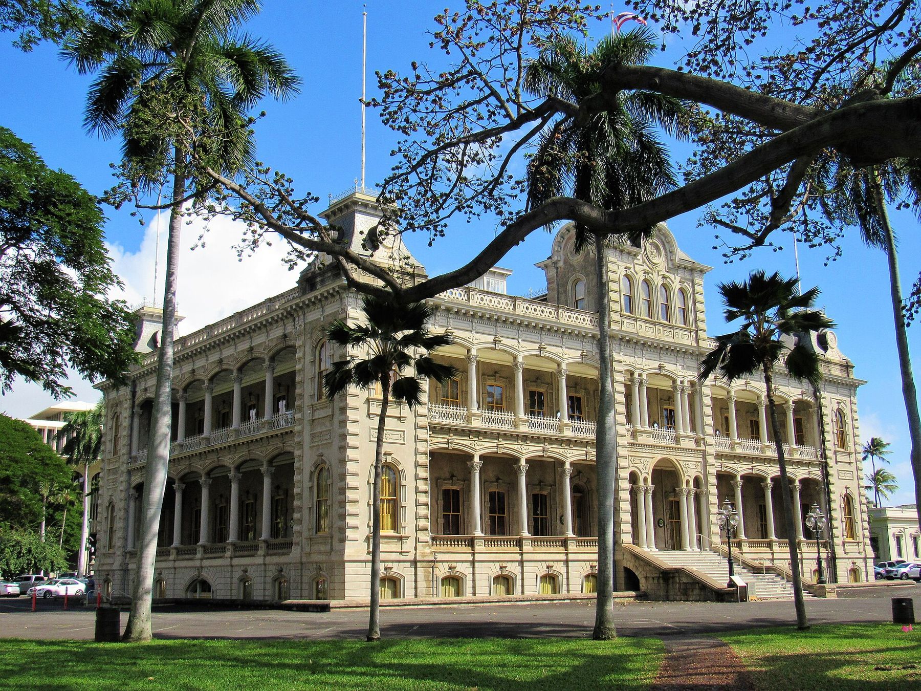 Iolani Palace in Honolulu, a grand historic building with arched balconies, framed by palm trees and a sprawling tree branch under a clear blue sky.