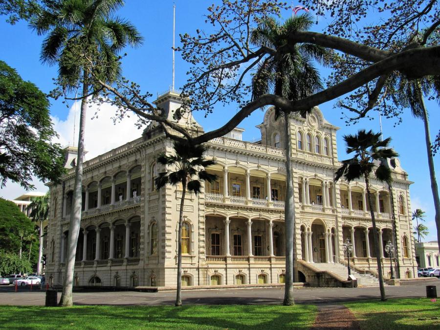 Iolani Palace in Honolulu, a grand historic building with arched balconies, framed by palm trees and a sprawling tree branch under a clear blue sky.
