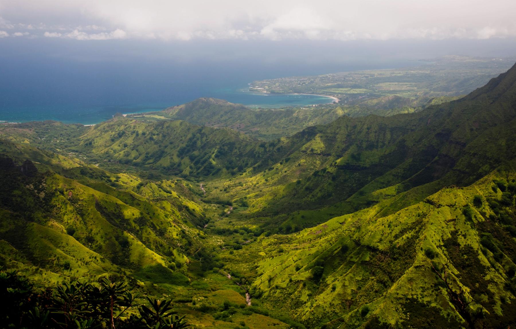 Sunlit green mountain ridges and a deep valley leading to the turquoise shoreline of Hanalei Bay under a hazy sky