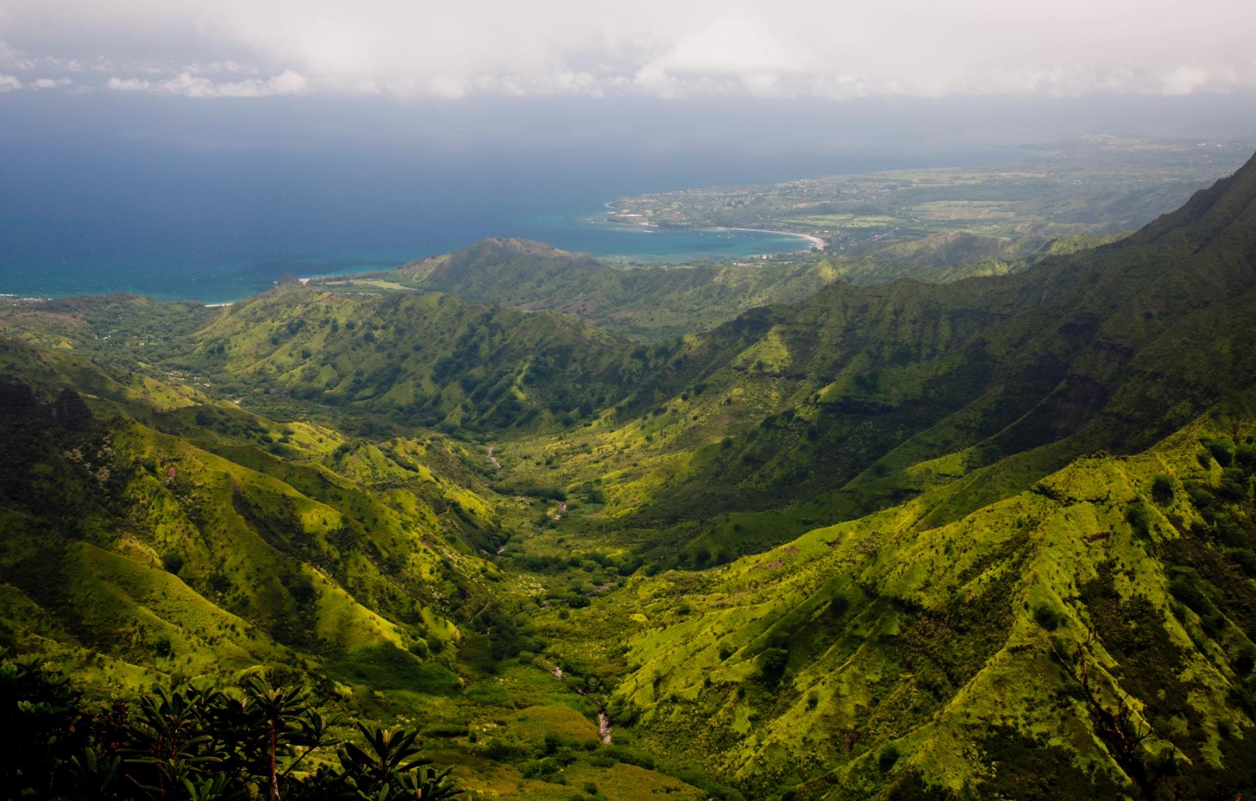 Sunlit green mountain ridges and a deep valley leading to the turquoise shoreline of Hanalei Bay under a hazy sky
