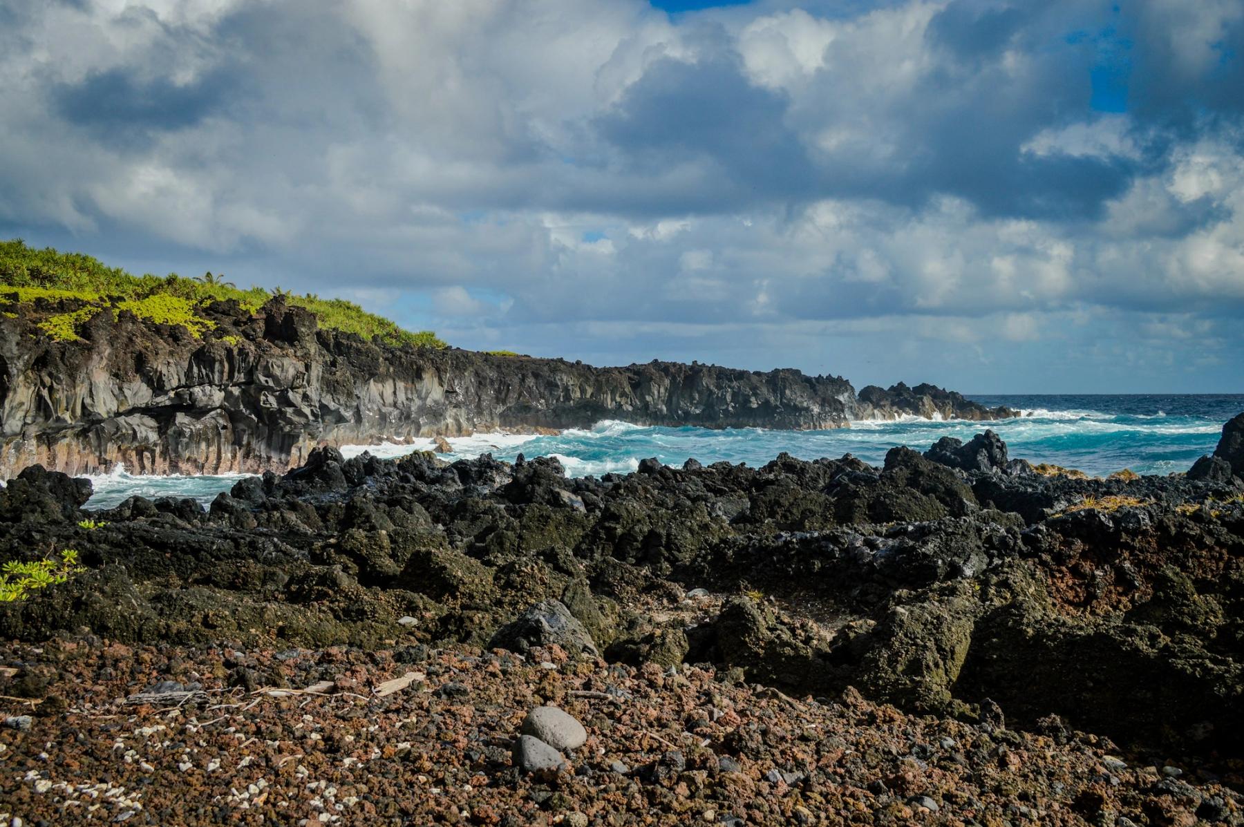 Rocky lava coastline in East Maui with waves breaking against black cliffs under a partly cloudy sky.