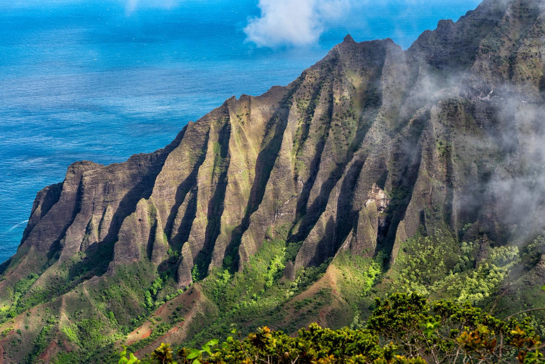 Sunlit Nā Pali Coast ridges with wisps of cloud over steep green valleys and deep blue ocean