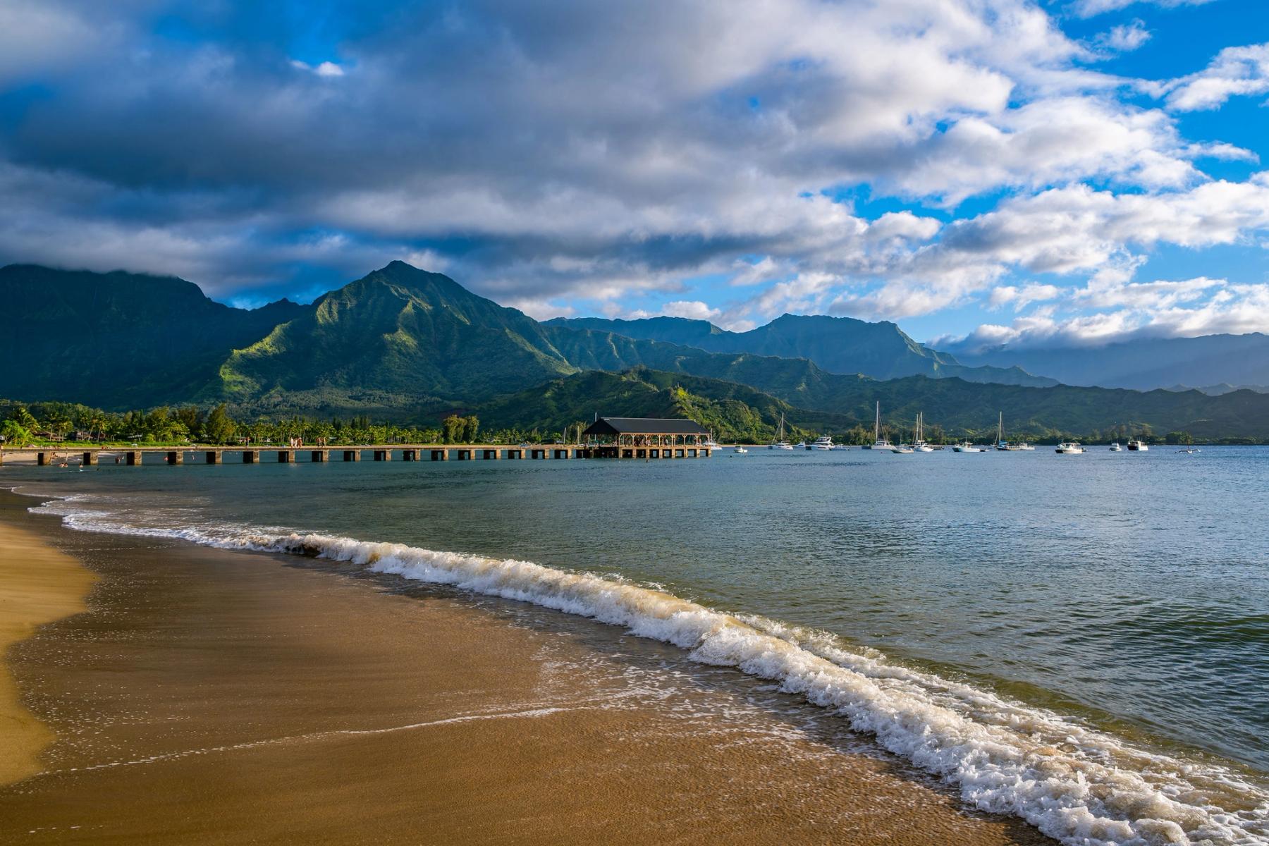 Curving sandy beach and small waves leading toward Hanalei Bay pier, with sailboats on calm water and green mountains beneath dramatic clouds