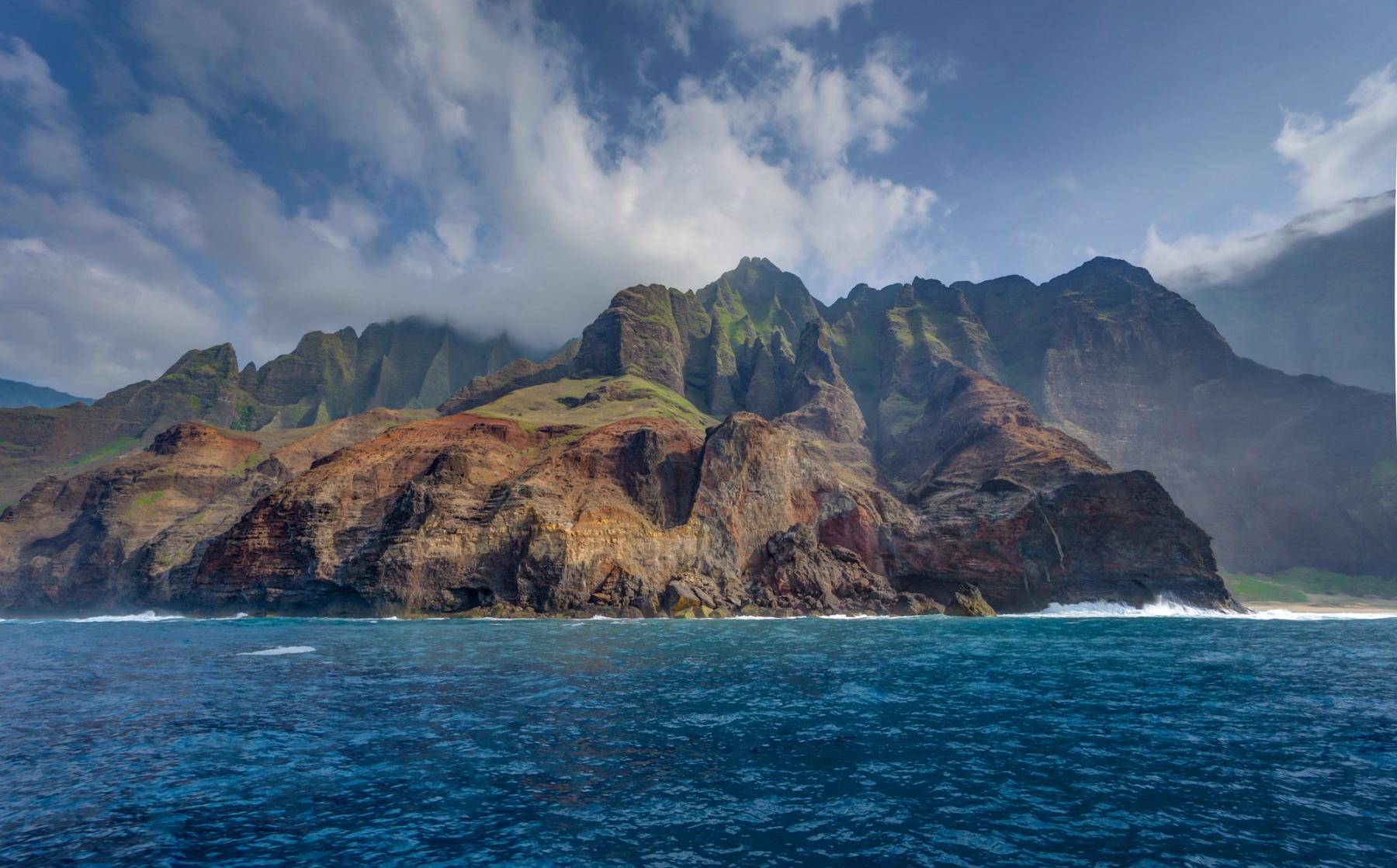 Towering Nā Pali Coast sea cliffs rising from deep blue ocean beneath sweeping white clouds