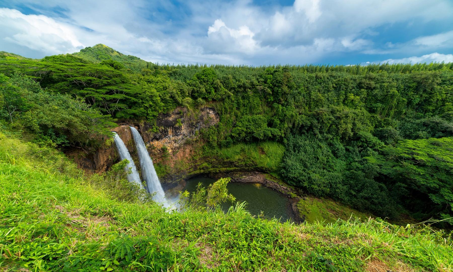 Wide view of Wailua Falls cascading into a circular pool amid dense green rainforest under a bright, cloud-filled sky