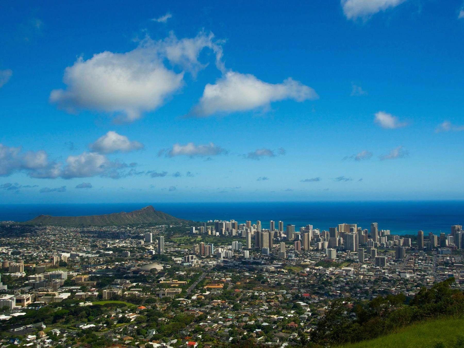 Panoramic view of Honolulu’s skyline and neighborhoods along the coastline, with Diamond Head in the distance under a blue sky with clouds.