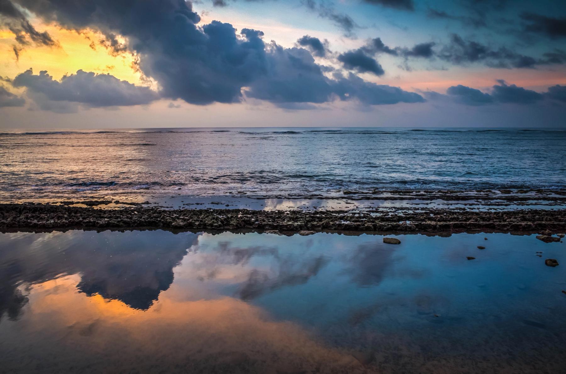 Sunrise glow behind dark clouds over the ocean, reflected in a calm tidepool with rocky shoreline in the foreground