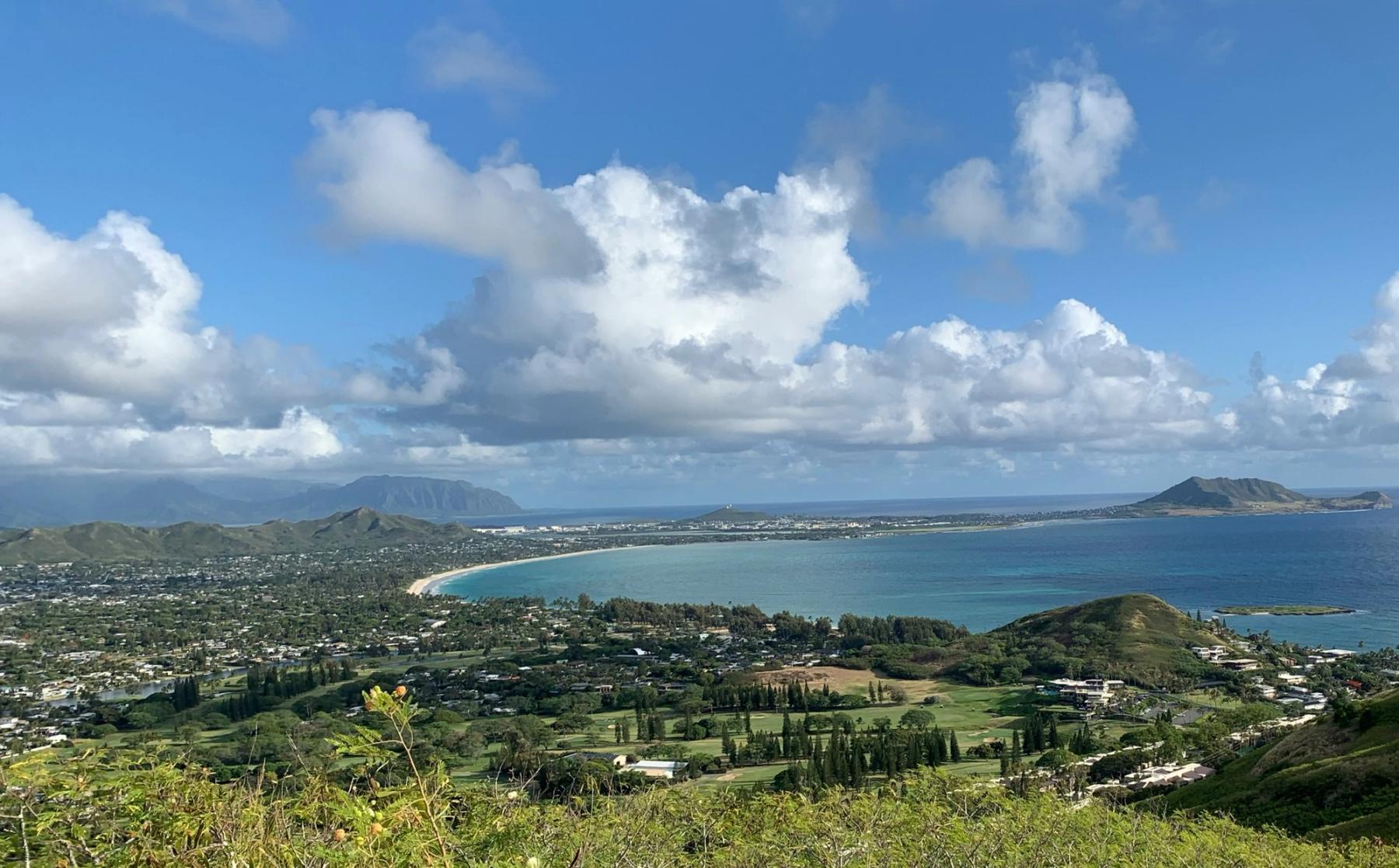 Elevated view of a crescent bay, coastal town, and green hills under scattered clouds on Oʻahu’s Windward Coast.