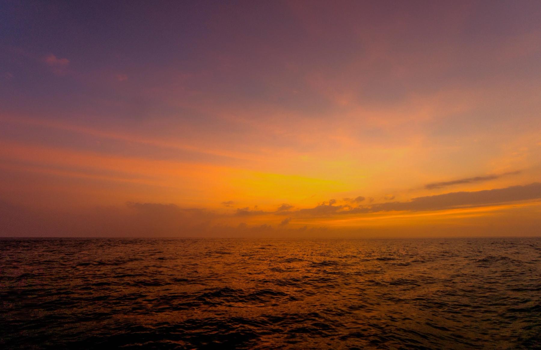Golden-orange sunset over the Pacific with a low horizon, layered clouds, and dark rippling water reflecting warm light