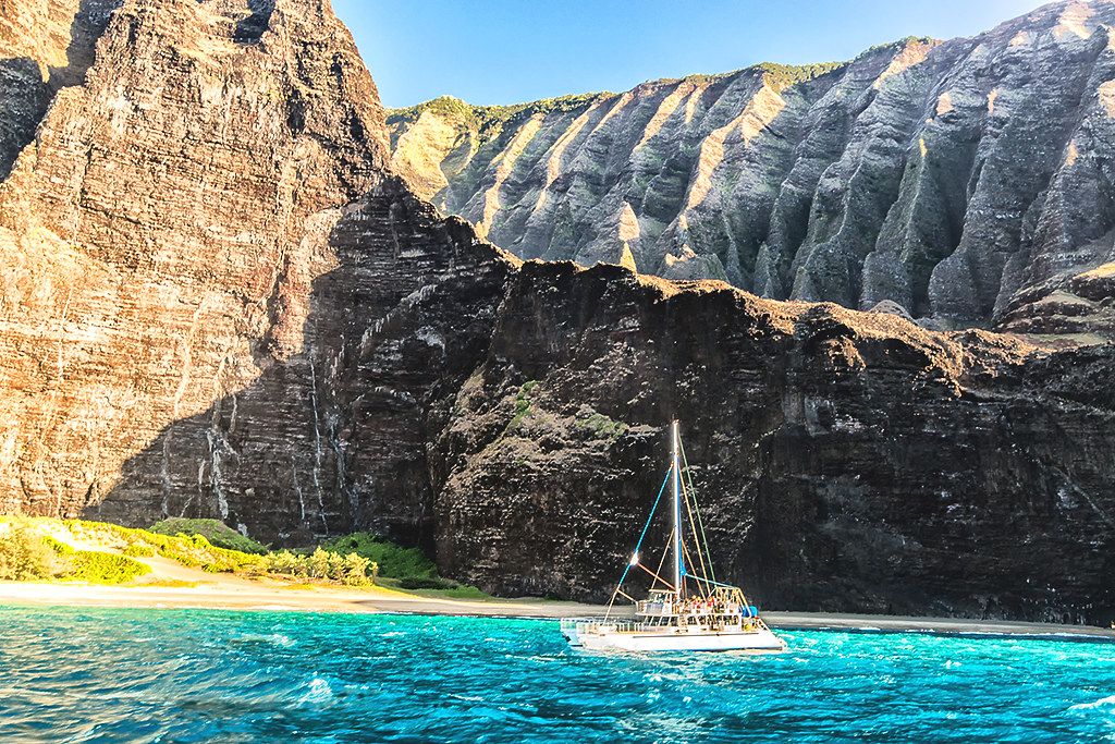 Sailboat in bright turquoise water below towering Nā Pali Coast cliffs and a small sandy beach
