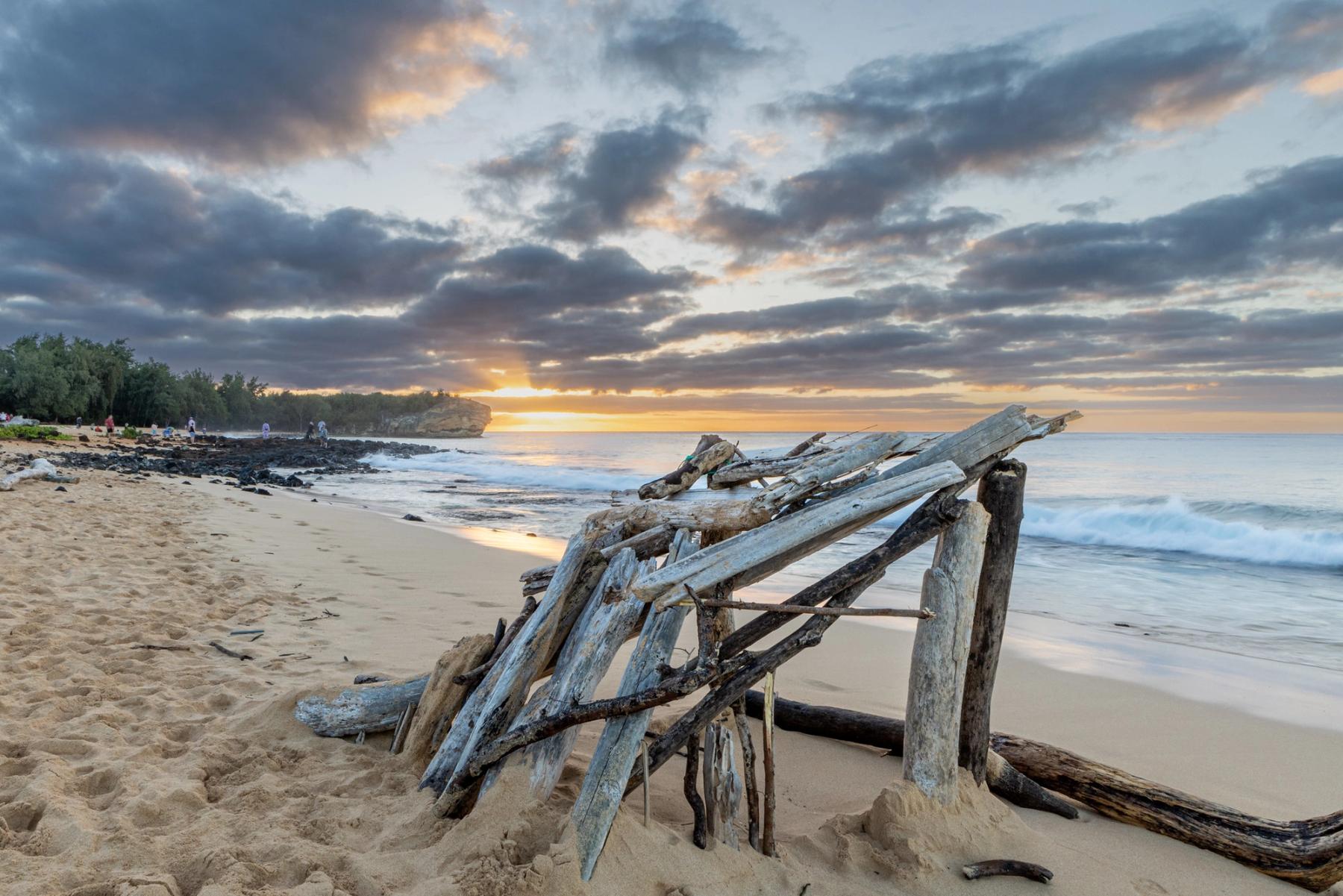Weathered driftwood structure on sandy beach with gentle surf and a glowing sunrise beneath dramatic clouds