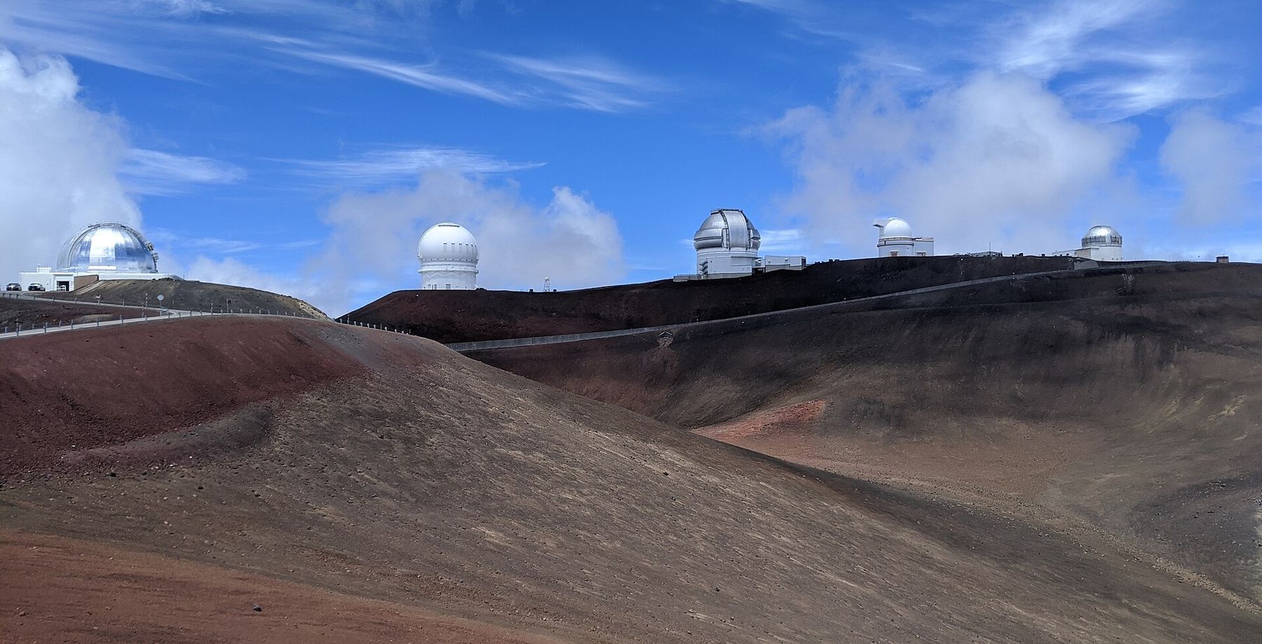 Observatory domes line a ridgeline above barren volcanic slopes under a bright blue sky at Mauna Kea Summit.