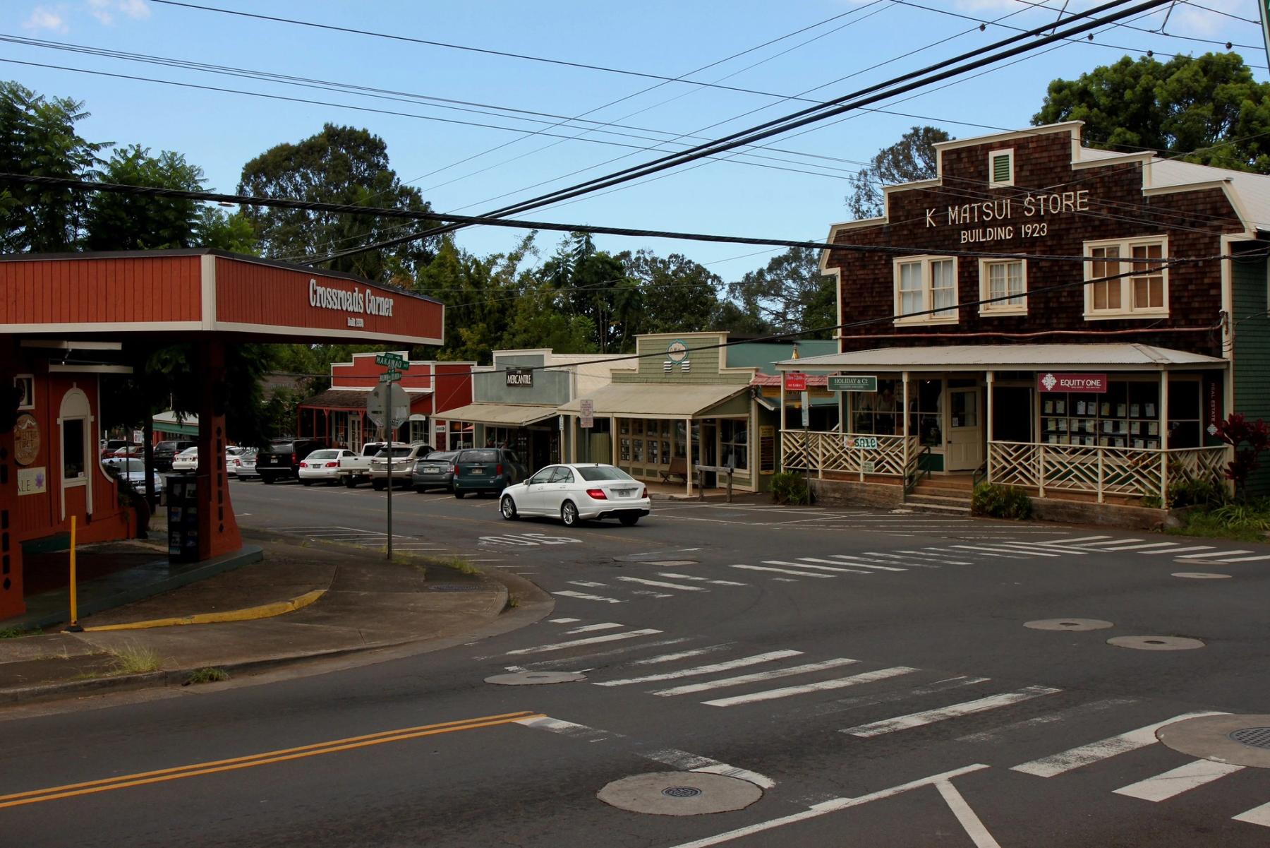 Street intersection in Makawao, Maui with storefronts, parked cars, a white car passing, and overhead power lines.