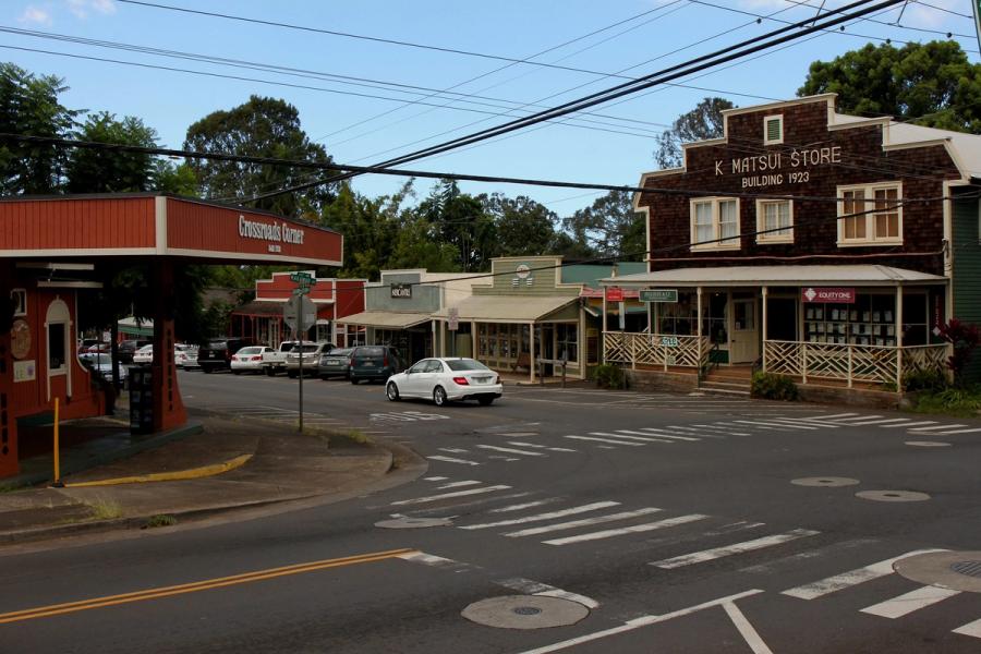 Street intersection in Makawao, Maui with storefronts, parked cars, a white car passing, and overhead power lines.