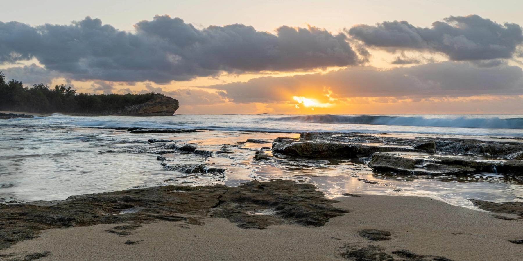 Sunrise over the ocean with layered clouds, waves, and wet lava-rock shelves and tide pools reflecting warm light in the foreground.