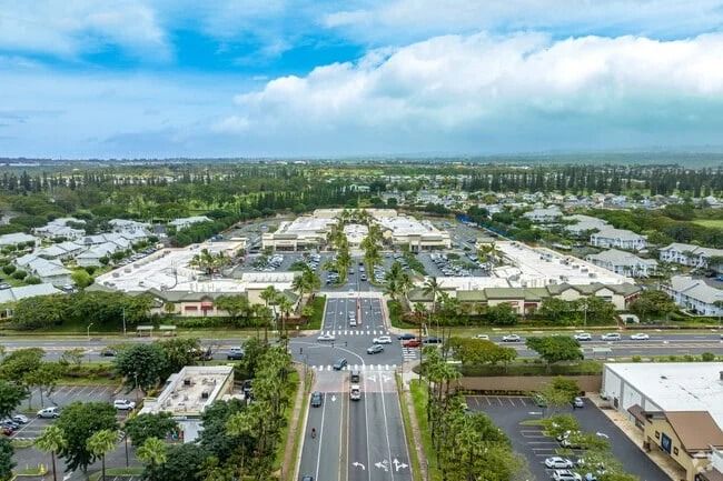 Aerial view of a shopping center and surrounding neighborhoods along a wide road in Waipahu, Oahu, with low clouds over green hills in the distance.