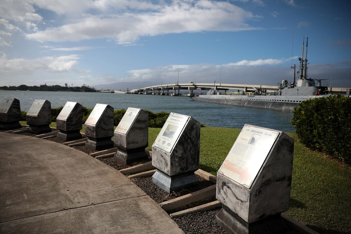 Stone informational plaques line a waterfront walkway with a submarine and bridge visible across the harbor at the Pearl Harbor National Memorial on Oʻahu.