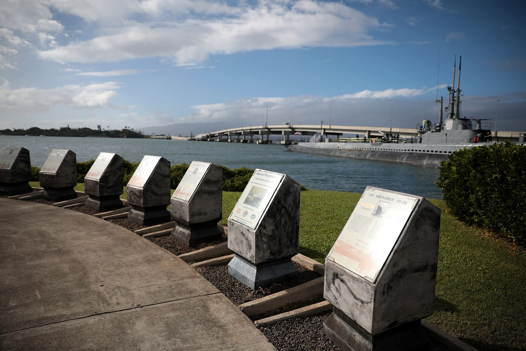 Stone informational plaques line a waterfront walkway with a submarine and bridge visible across the harbor at the Pearl Harbor National Memorial on Oʻahu.