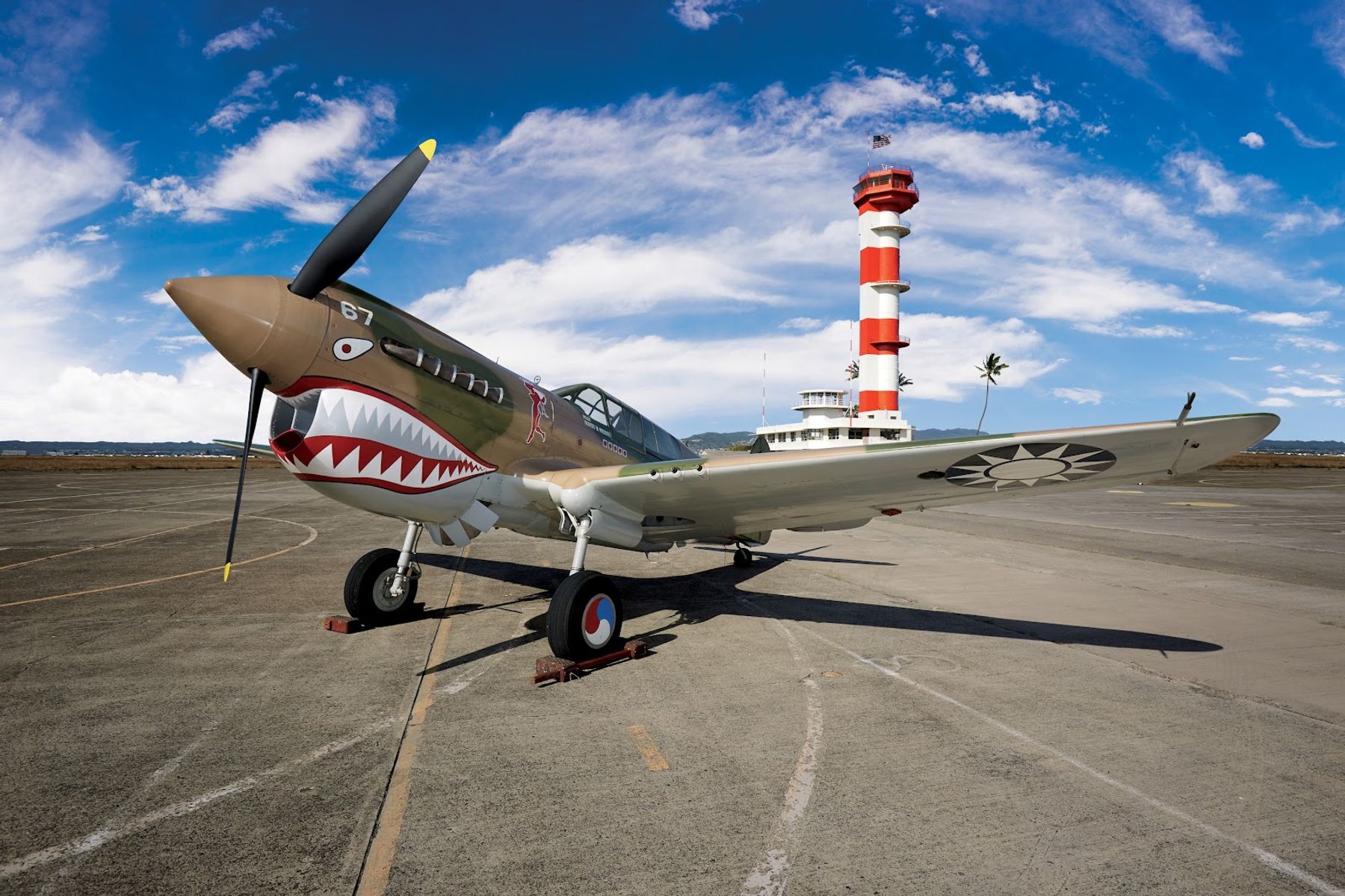 A shark-mouth fighter aircraft displayed outside Pearl Harbor Aviation Museum with the red-and-white Ford Island Control Tower behind it.