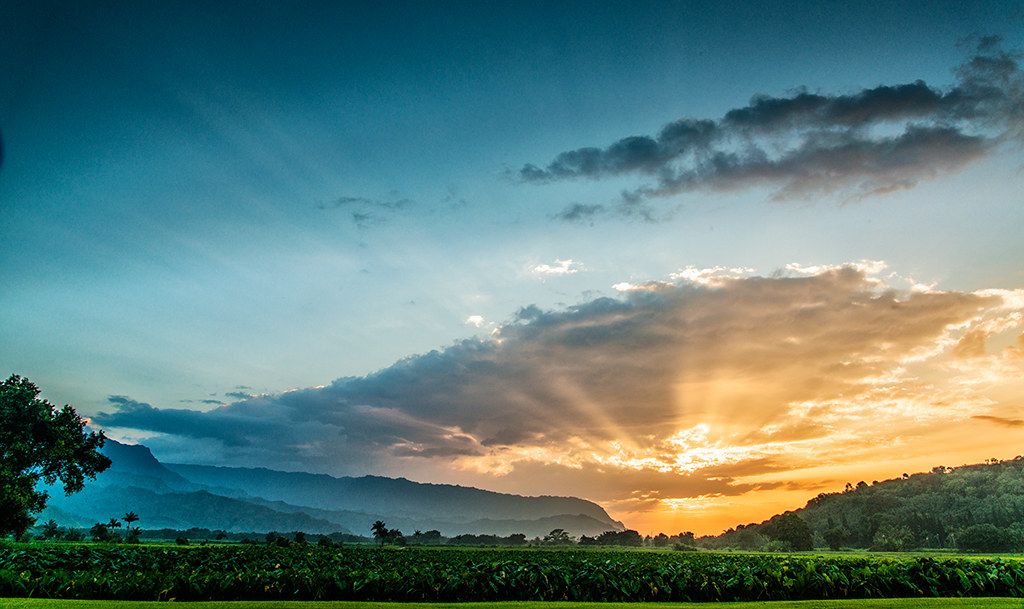 Sunset rays breaking through clouds over Hanalei Valley mountains with green taro fields in the foreground on Kauaʻi.