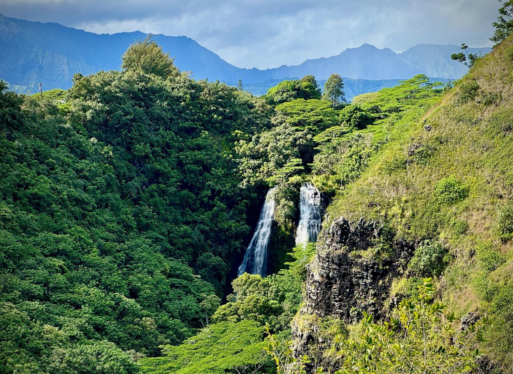 Twin waterfalls cascading into a lush green Kauaʻi valley with rugged mountain ridges under a hazy sky