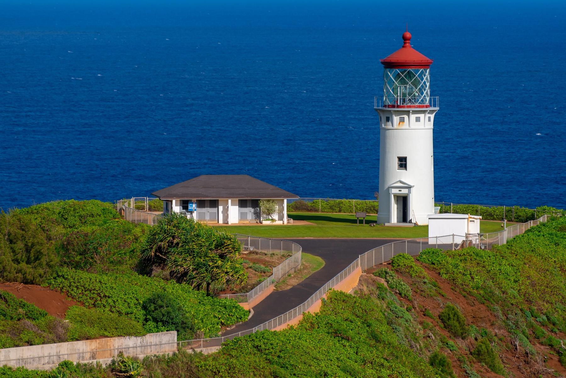 White lighthouse with a red roof on a green coastal point, curving road and buildings in the foreground, deep blue ocean behind