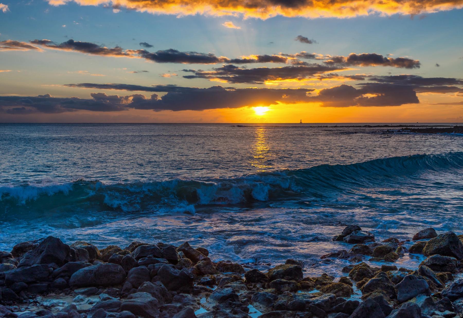 Sunset over the ocean with glowing clouds, a bright reflection on the water, a curling wave, and dark lava rocks in the foreground