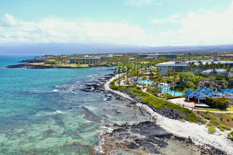 Aerial view of Waikoloa coastline on Hawaiʻi Island with resort buildings, palm trees, pools, and a rocky shore beside clear blue water.