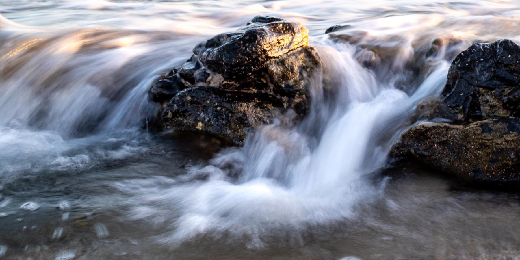 Long-exposure water flowing over dark volcanic rocks at the shoreline with a warm sunlit highlight on the top rock