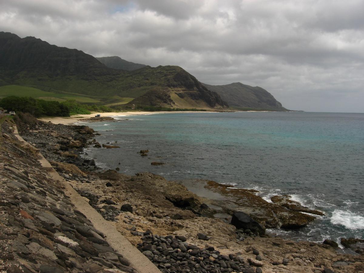 Rocky shoreline and a narrow beach beneath green mountains on Oʻahu’s Leeward Coast under a cloudy sky.
