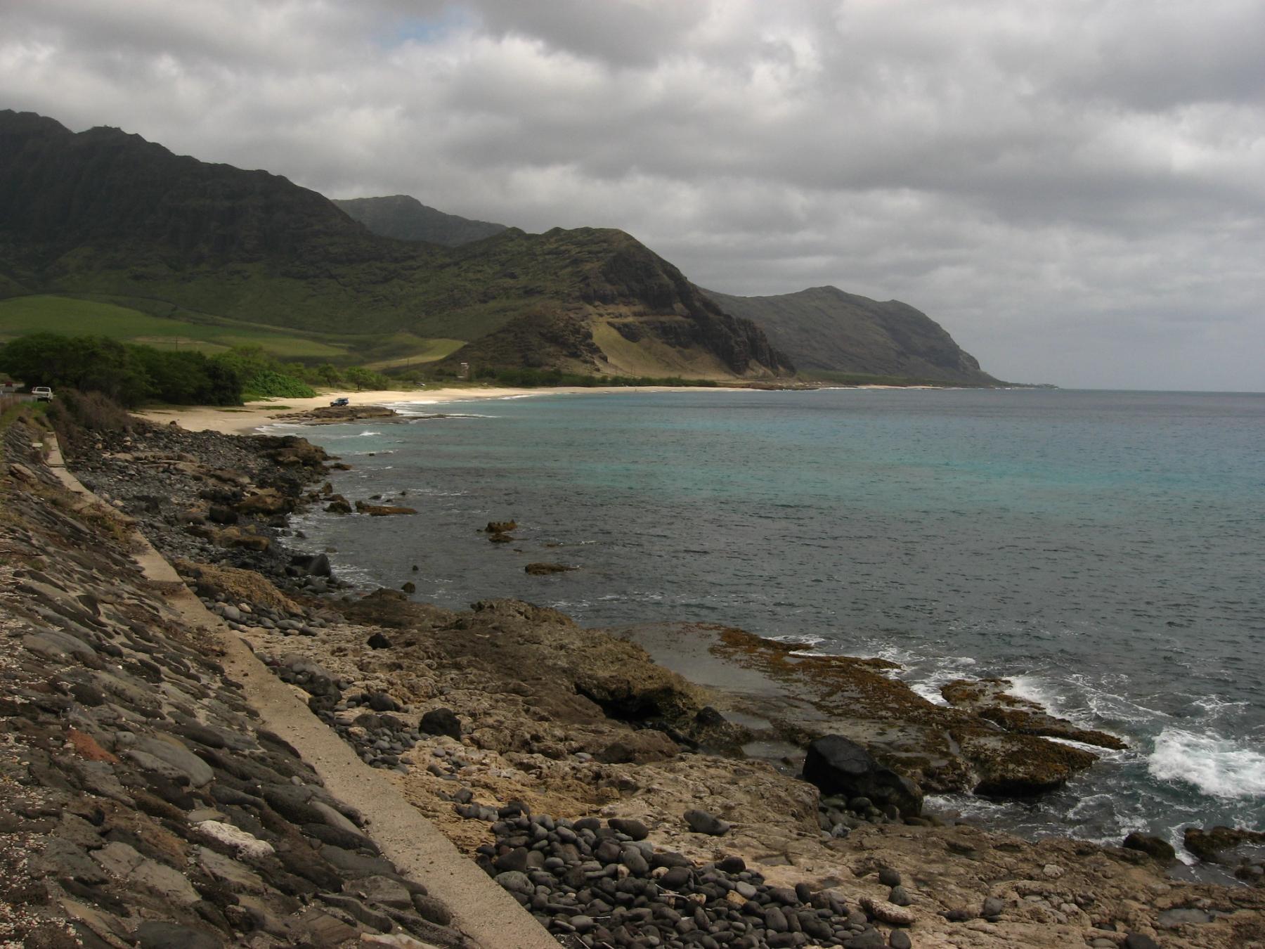 Rocky shoreline and a narrow beach beneath green mountains on Oʻahu’s Leeward Coast under a cloudy sky.