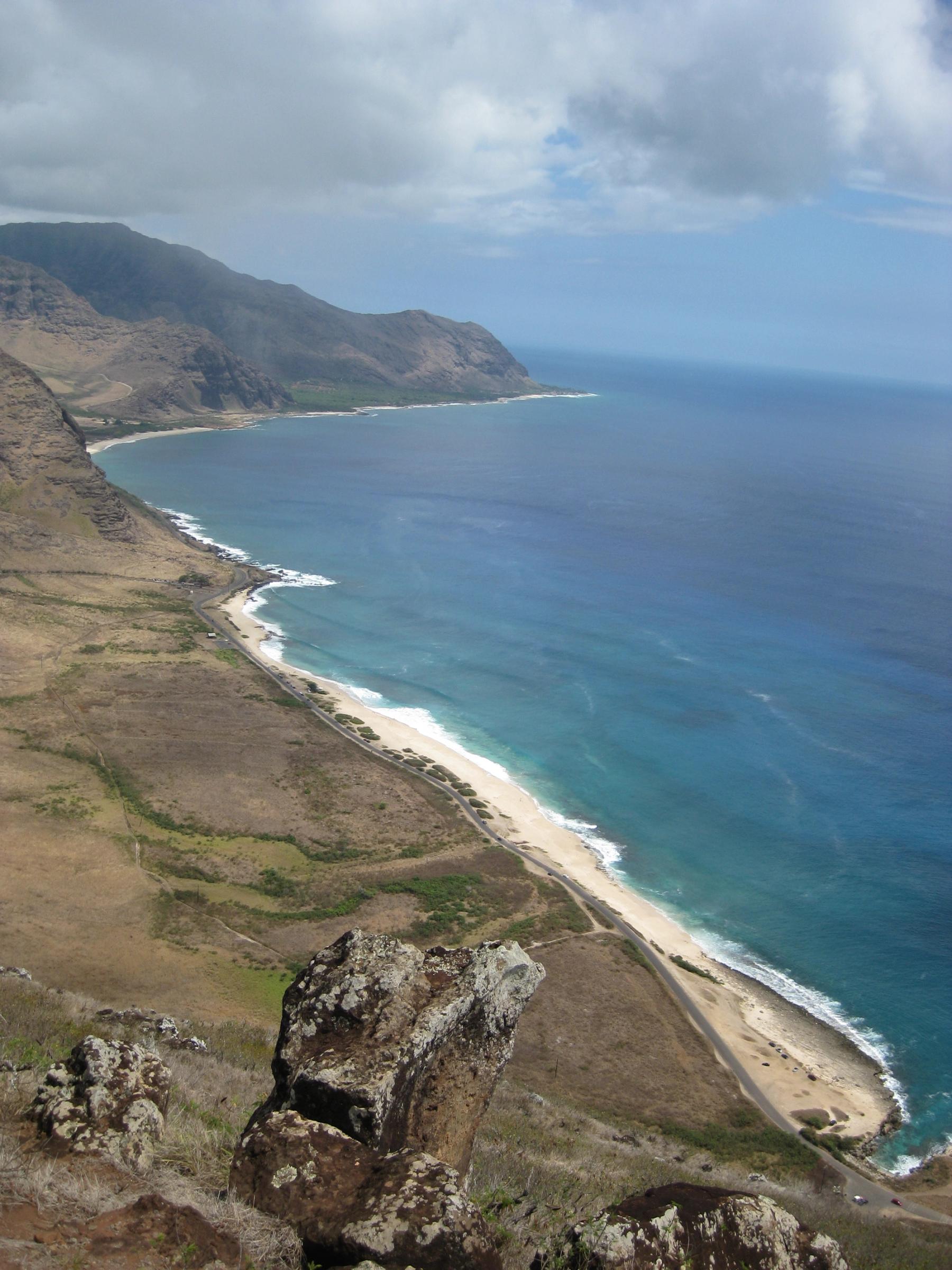 View from a rocky cliff over a long sandy shoreline and coastal road with rugged mountains beside the ocean on Oʻahu’s Waiʻanae Coast.