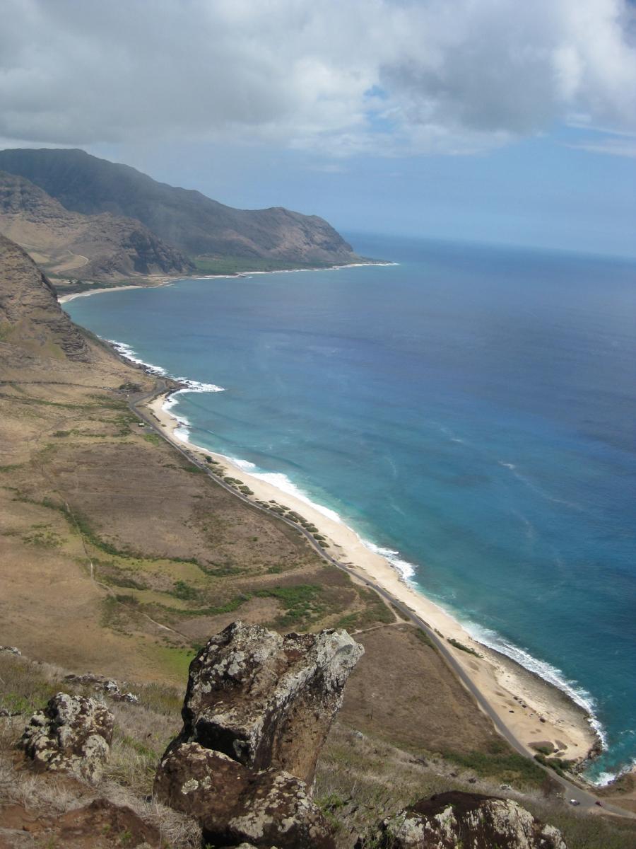 View from a rocky cliff over a long sandy shoreline and coastal road with rugged mountains beside the ocean on Oʻahu’s Waiʻanae Coast.