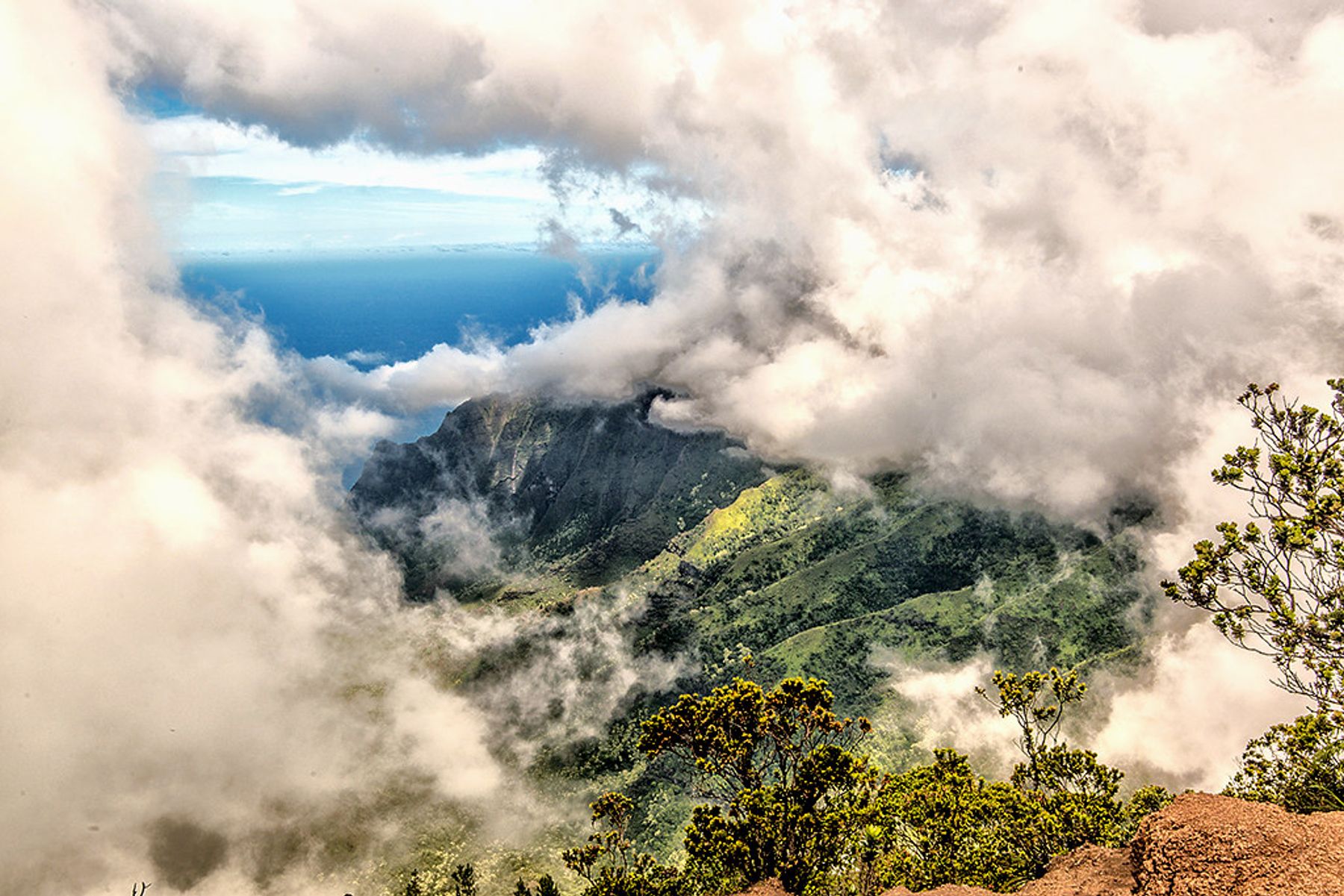 Clouds swirl around steep green ridges and valleys with the blue ocean visible beyond from a high Waimea Canyon lookout.